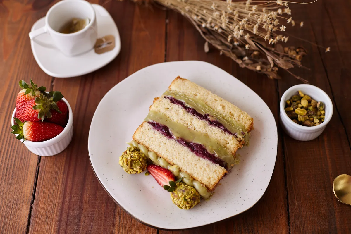 Fatia de bolo de pistache com frutas vermelhas servida com chá, morangos e pistaches em mesa de madeira